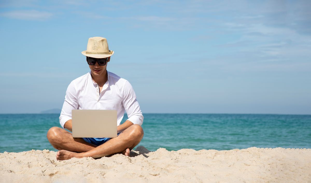 Asian man with a laptop doing work on the beach.