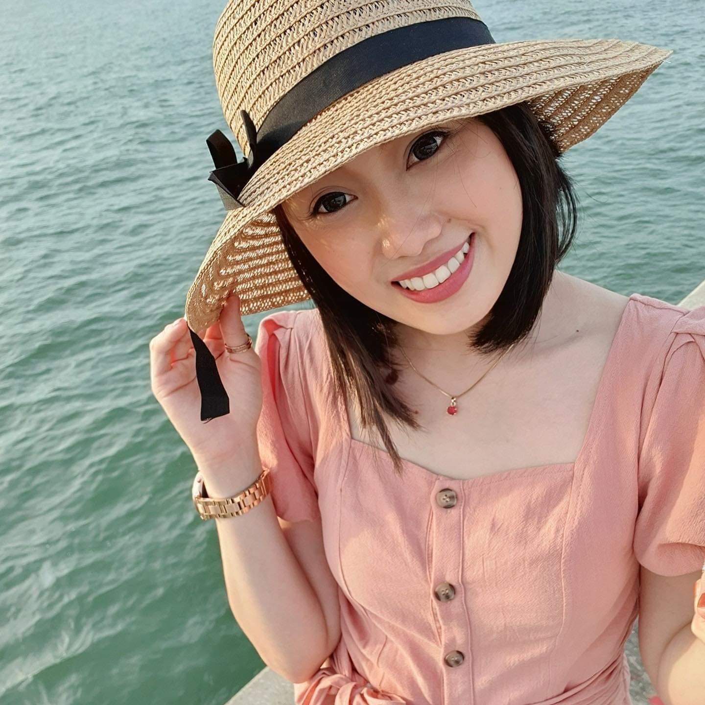 Asian woman wearing a hat and a peach dress posing by the sea. 