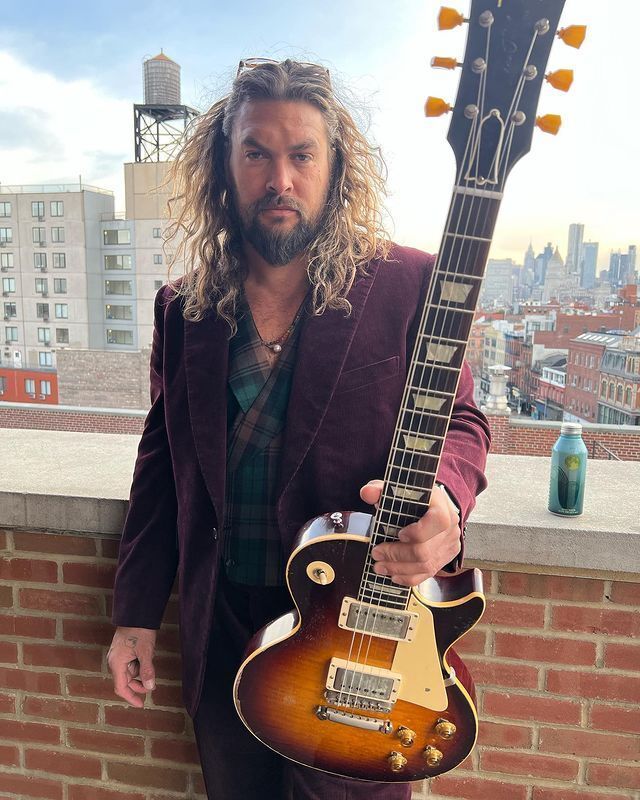 Jason Momoa with slicked back long hair holding a guitar.