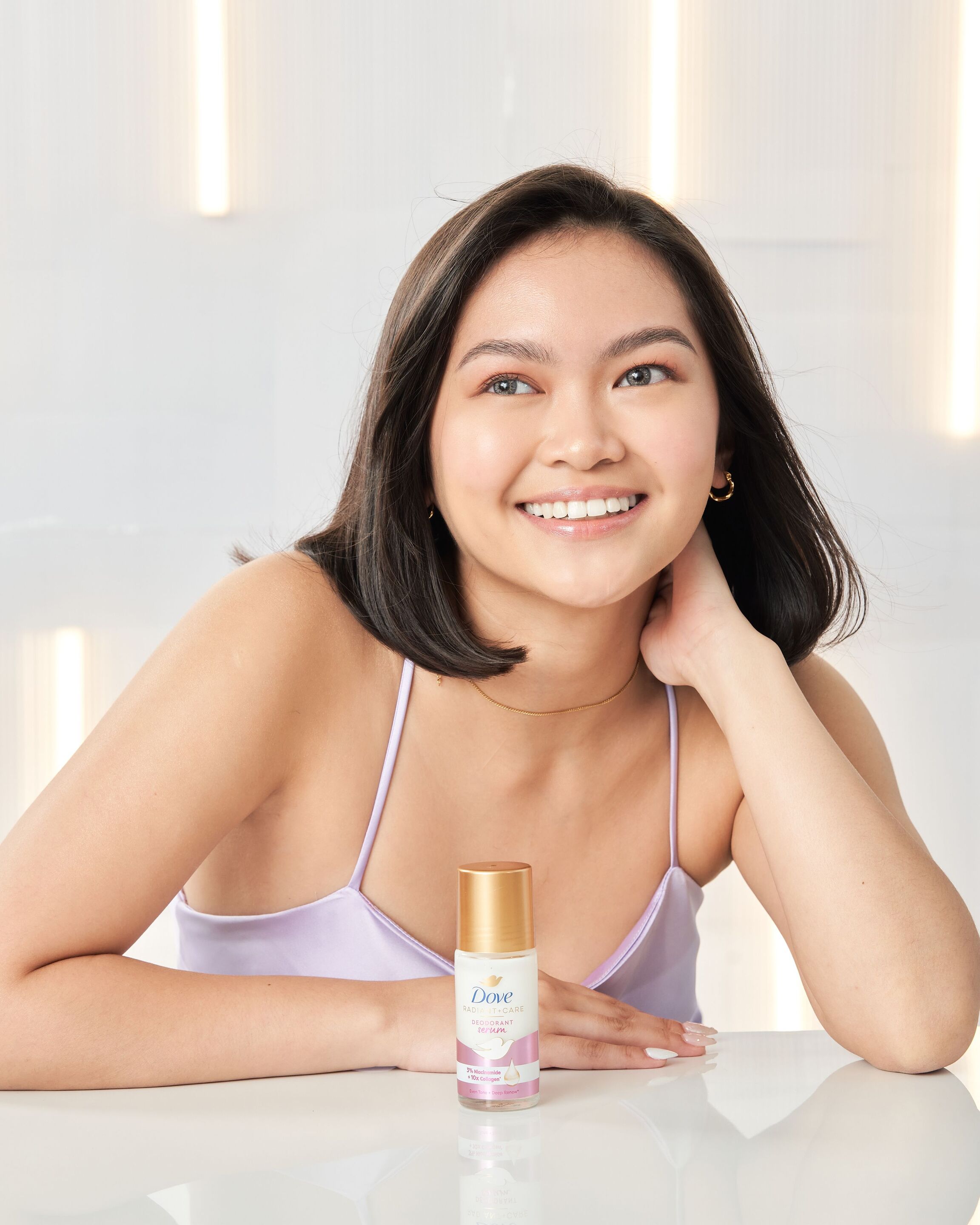 Woman in purple dress smiling with Dove Radiant + Care 3% Niacinamide + 10x Collagen Deep Renew Rollon on the table. 