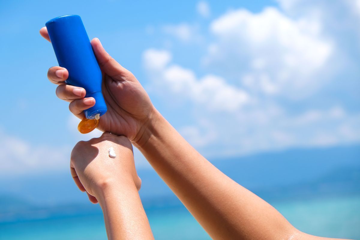 Closeup of woman holding a blue bottle with one hand and applying sunscreen to her other hand against a background of the sea.