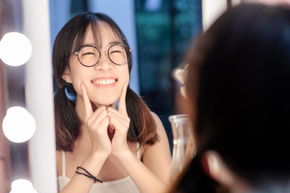 Woman with glasses happily checking her skin in the mirror.