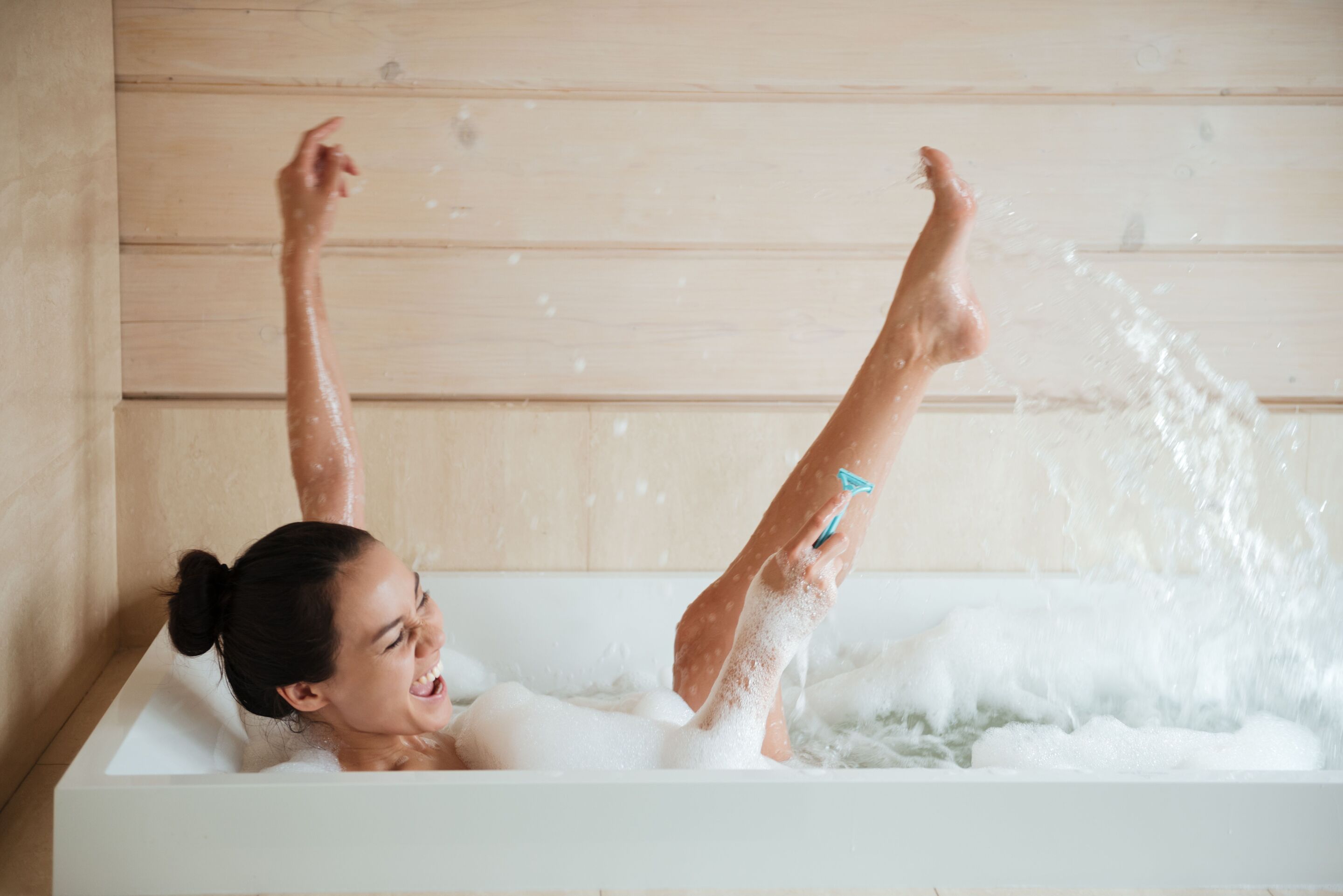 Asian woman in a hair bun laughing while taking a bath and shaving her legs