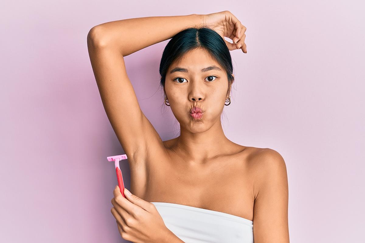 Asian woman with pursed lips, arm raised and holding pink razor