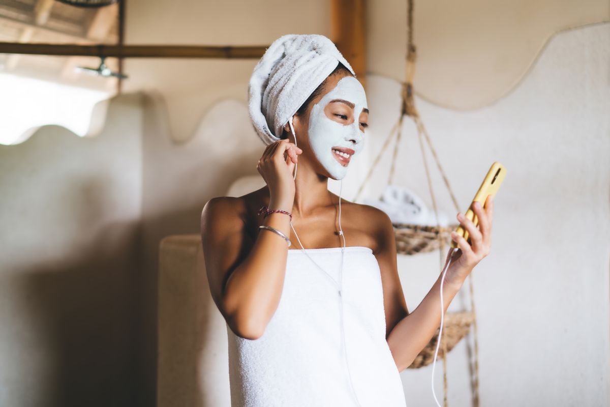 Asian woman with a clay mask on her face, listening to music on her phone.