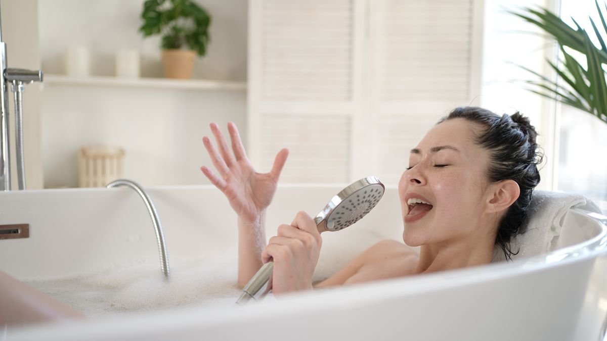 Woman in a bathtub singing with a shower head in her hand.