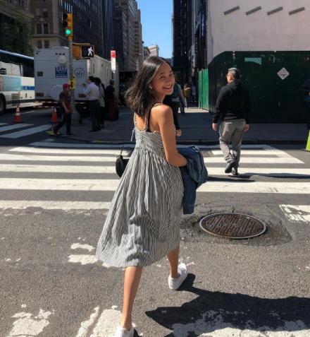 A woman in gray sundress walking on a zebra crossing and smiling at the camera behind