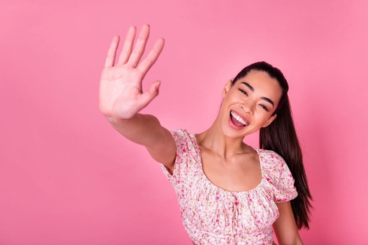 Asian woman smiling and holding her hand up against a pink background.
