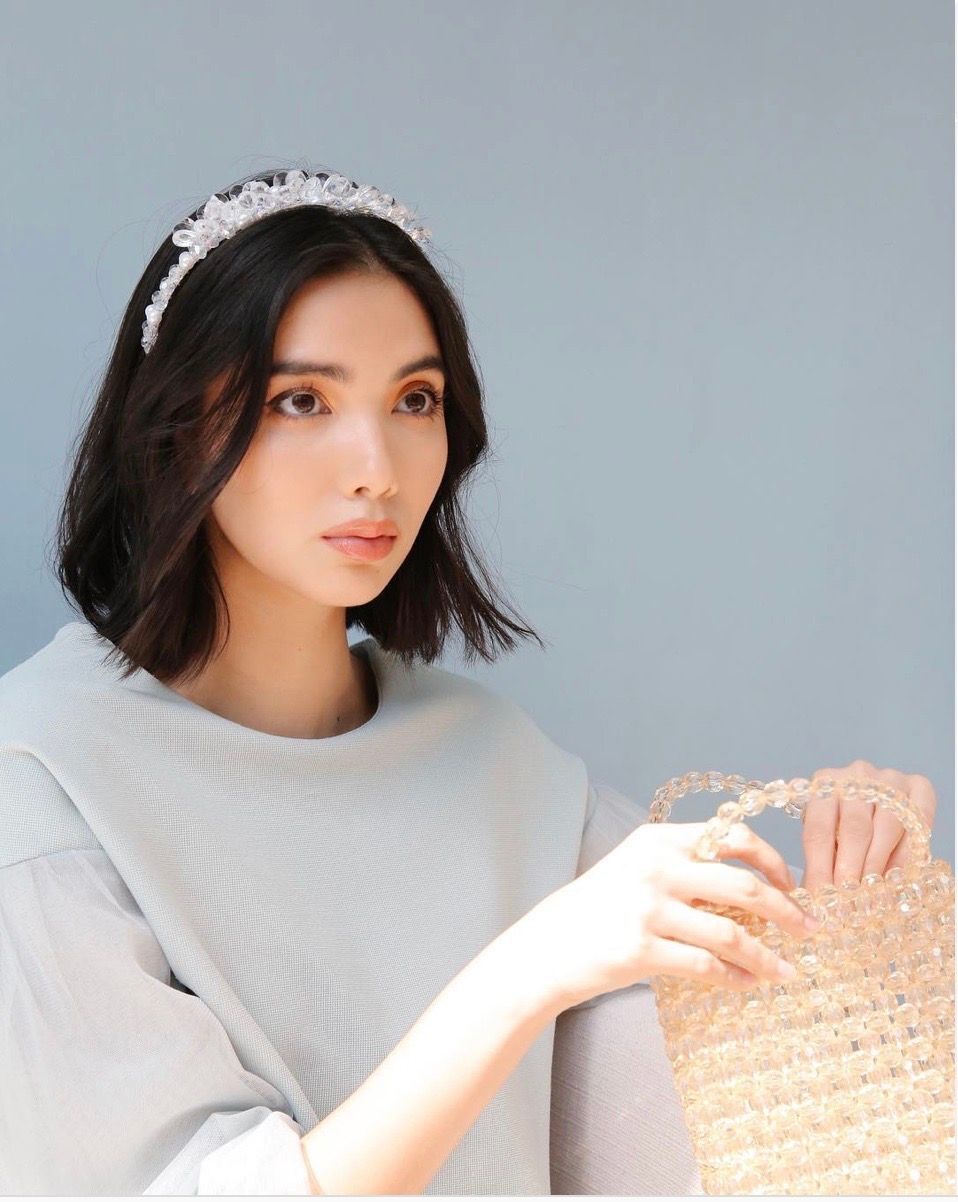 Asian woman with a bejeweled headband and bag against a grey background.