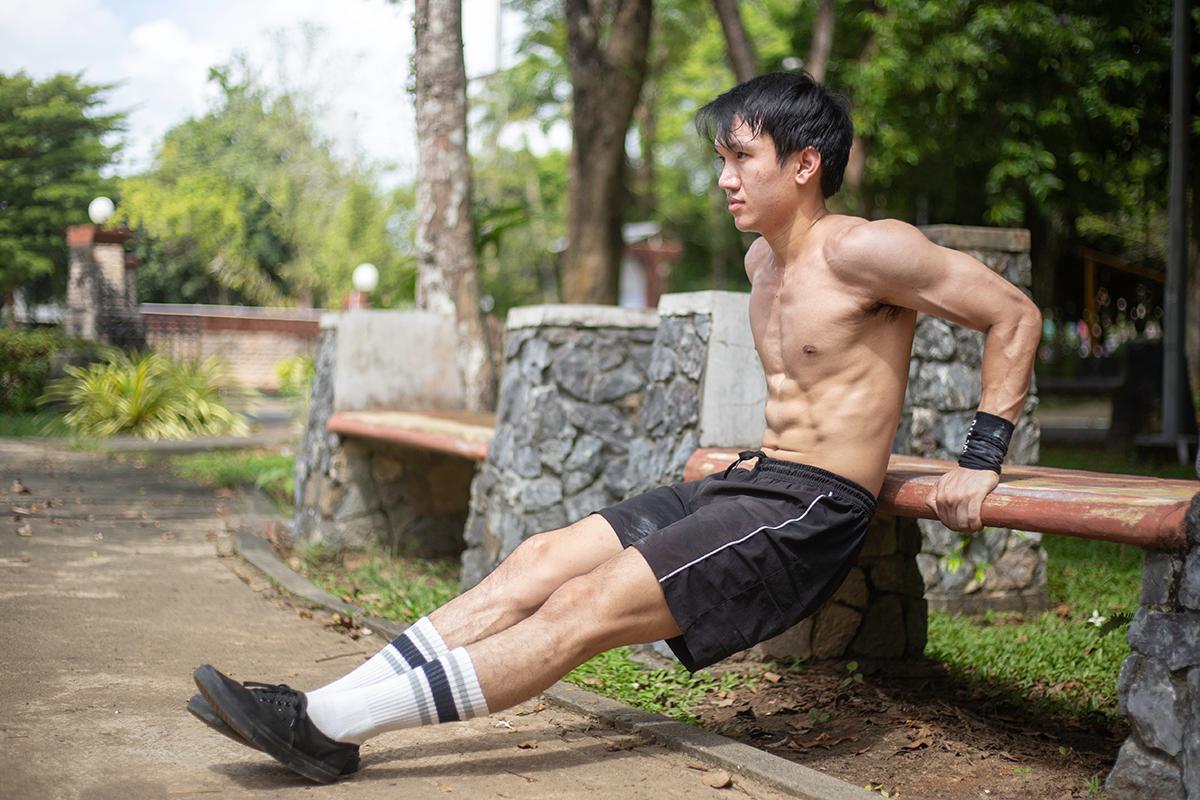 Young Asian man does tricep dips at an outdoor bench.