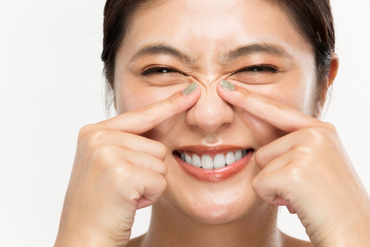 Asian woman smiling and crinkling her nose while pointing to it with her pointer fingers against a white background.