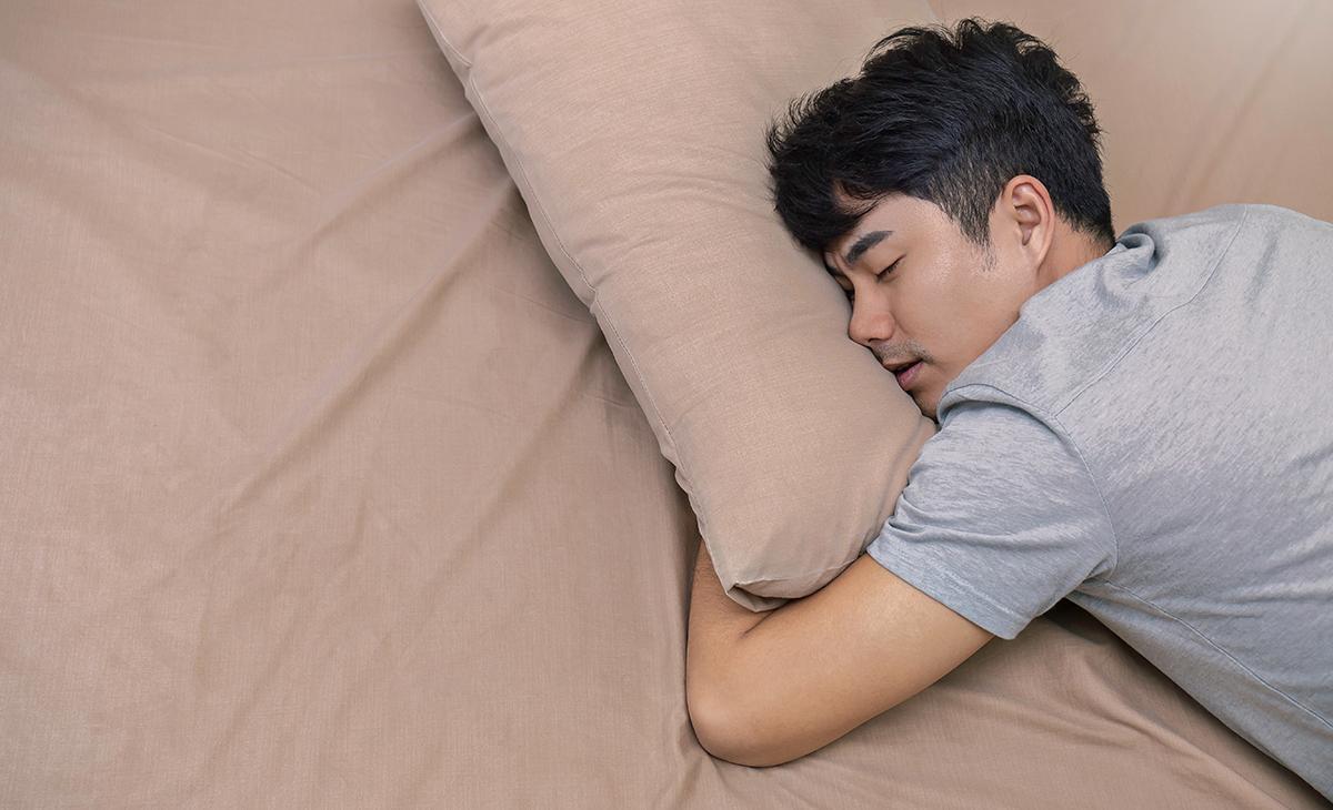 Young Asian man in gray shirt laying on brown sheets and hugging brown pillow 