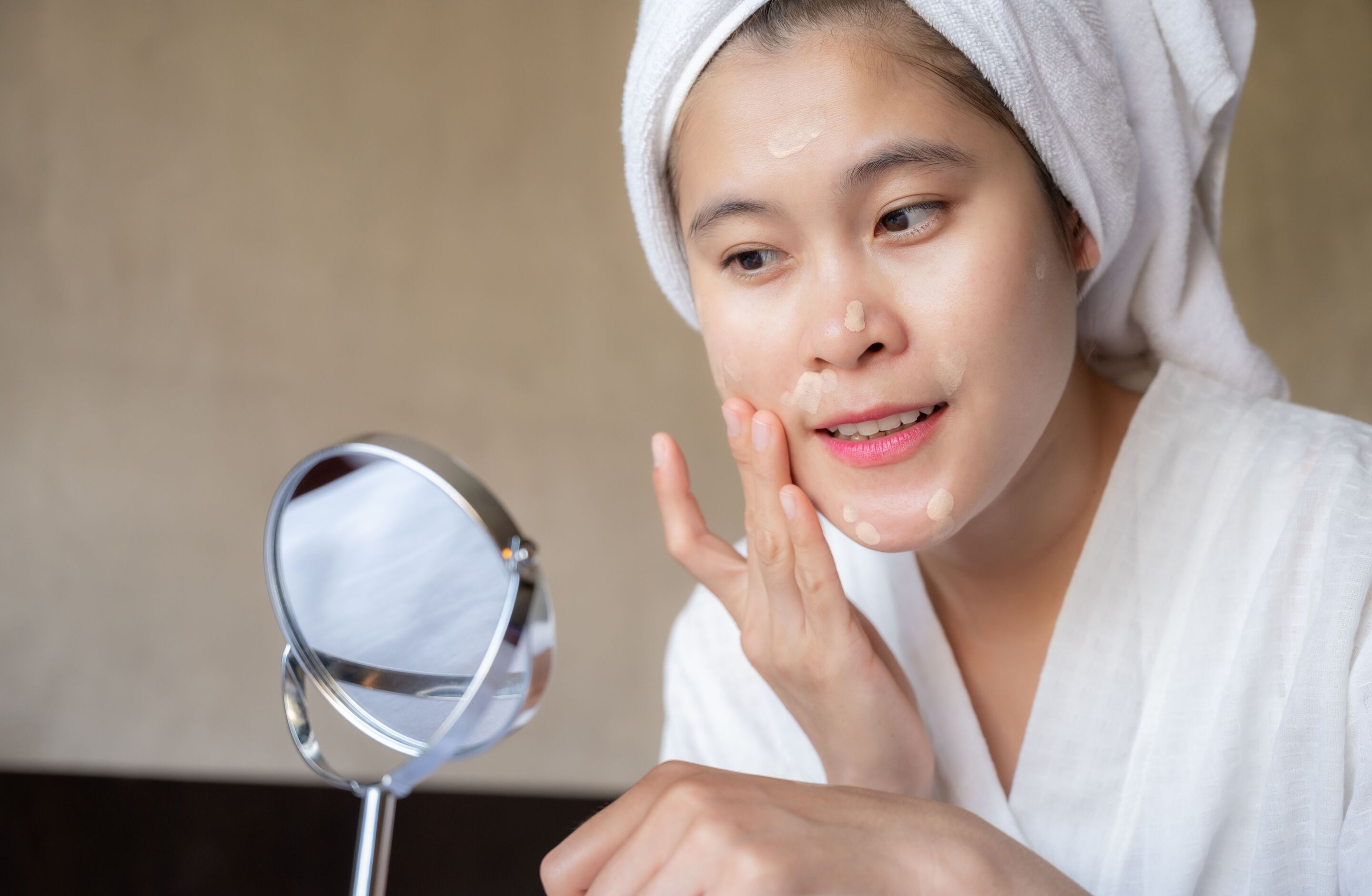 Woman in bathrobe applying liquid foundation on her face with her fingers.