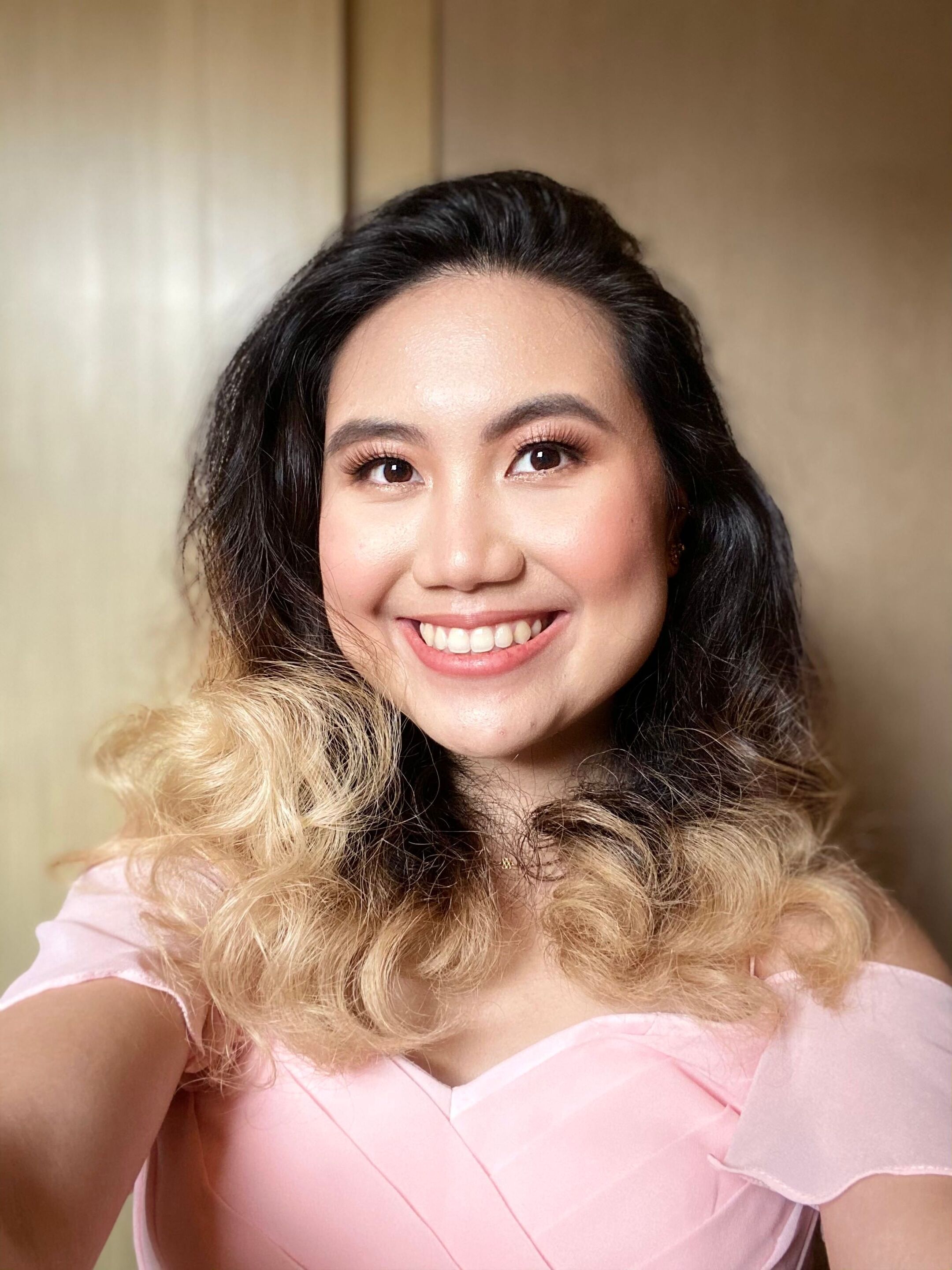 A portrait of a woman with blonde ombre hair wearing pink sabrina top.