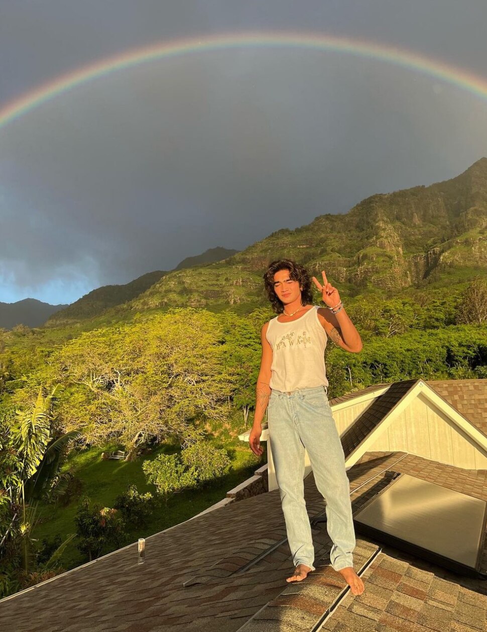 Bretman Rock on a roof making a peace sign with a rainbow behind him. 