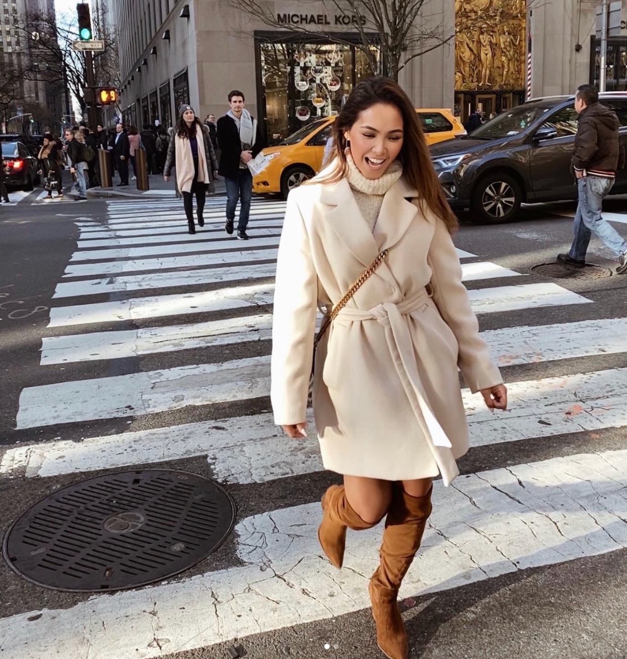 Woman wearing a white coat and suede boots smiling and walking down a crosswalk.