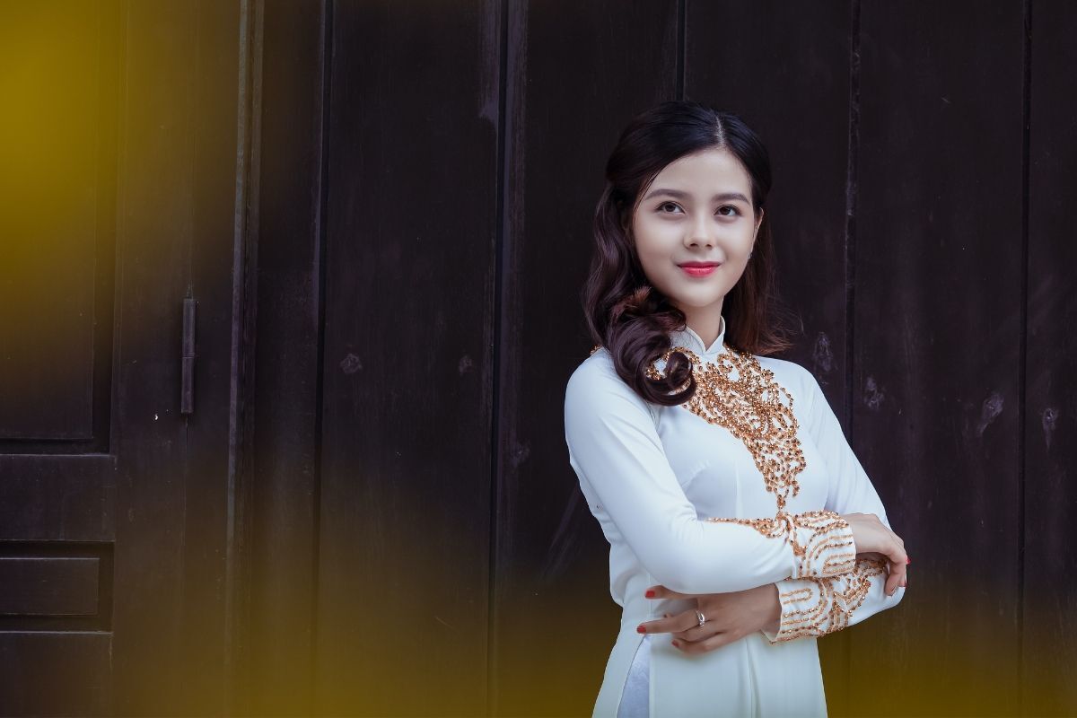 kiểu tóc mặc áo dài truyền thống cô gái mặc áo dài trắng có hoa văn màu vàng ánh kim tóc xoăn đuôi lọn to đứng khoanh tay