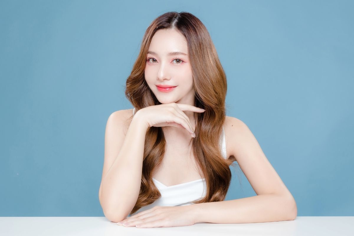 Cách trang điểm tự nhiên đơn giản tại nhà cho người mới bắt đầu cô gái chống tay vào cằm phía sau là nền tường màu xanh dương