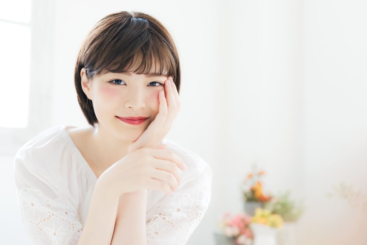 Tóc ngắn cho mặt tròn trán ngắn cô gái đặt tay lên má ảnh chụp trong phòng nhiều ánh sáng trắng phía sau là bình hoa hồng vàng