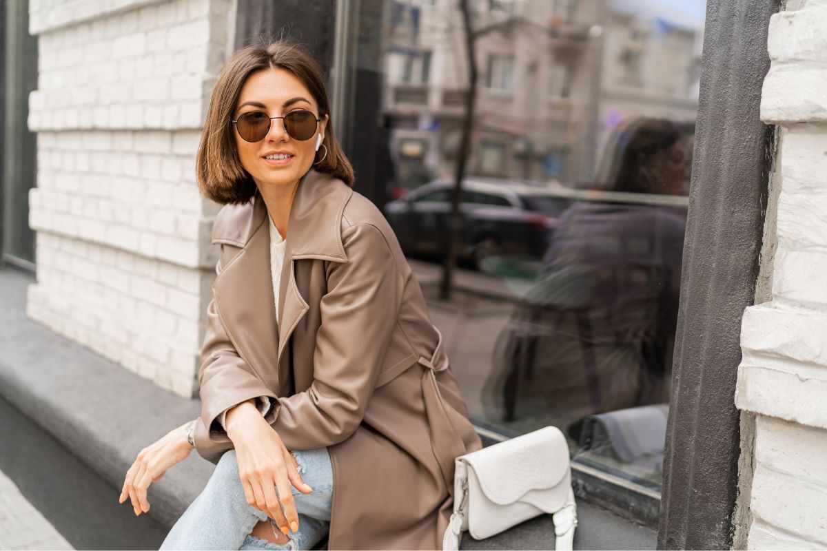 tóc ngắn uốn cụp cô gái mặc áo choàng dài màu nâu đeo kính râm bên cạnh là túi xách màu trắng