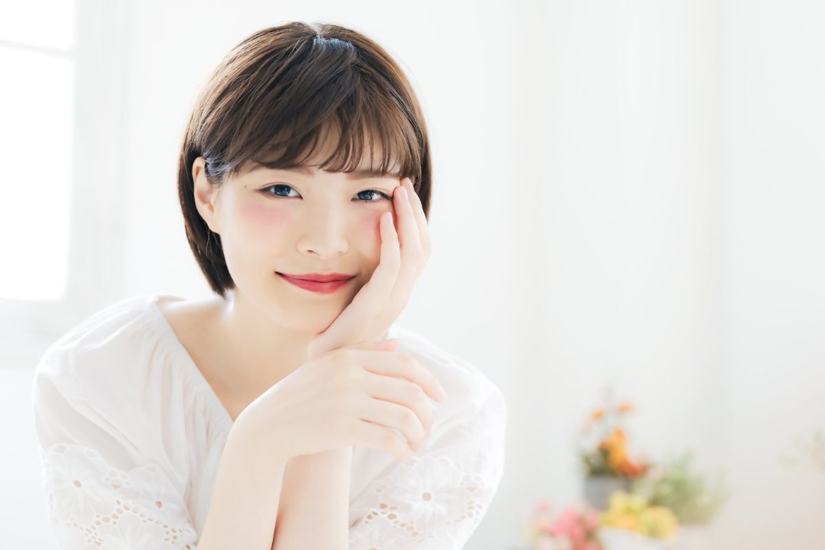 Tóc mái ngố cô gái mặc màu trắng đang áp một tay lên má phía sau là rèm cửa màu trắng và lọ hoa giả bị làm mờ