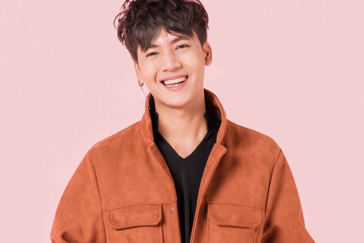 Kiểu tóc nam Hàn Quốc đẹp chàng trai mặc áo khoác màu cam đang cười tươi chụp hình trên phông nền màu hồng