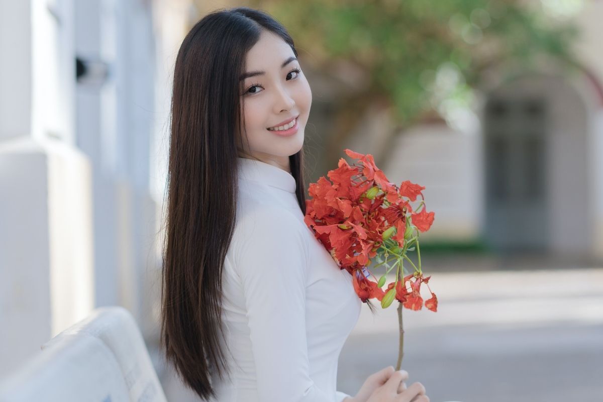 Cô gái tóc dài thẳng tay đang cầm nhành hoa phượng đỏ và tạo dáng chụp ảnh áo dài kỉ yếu