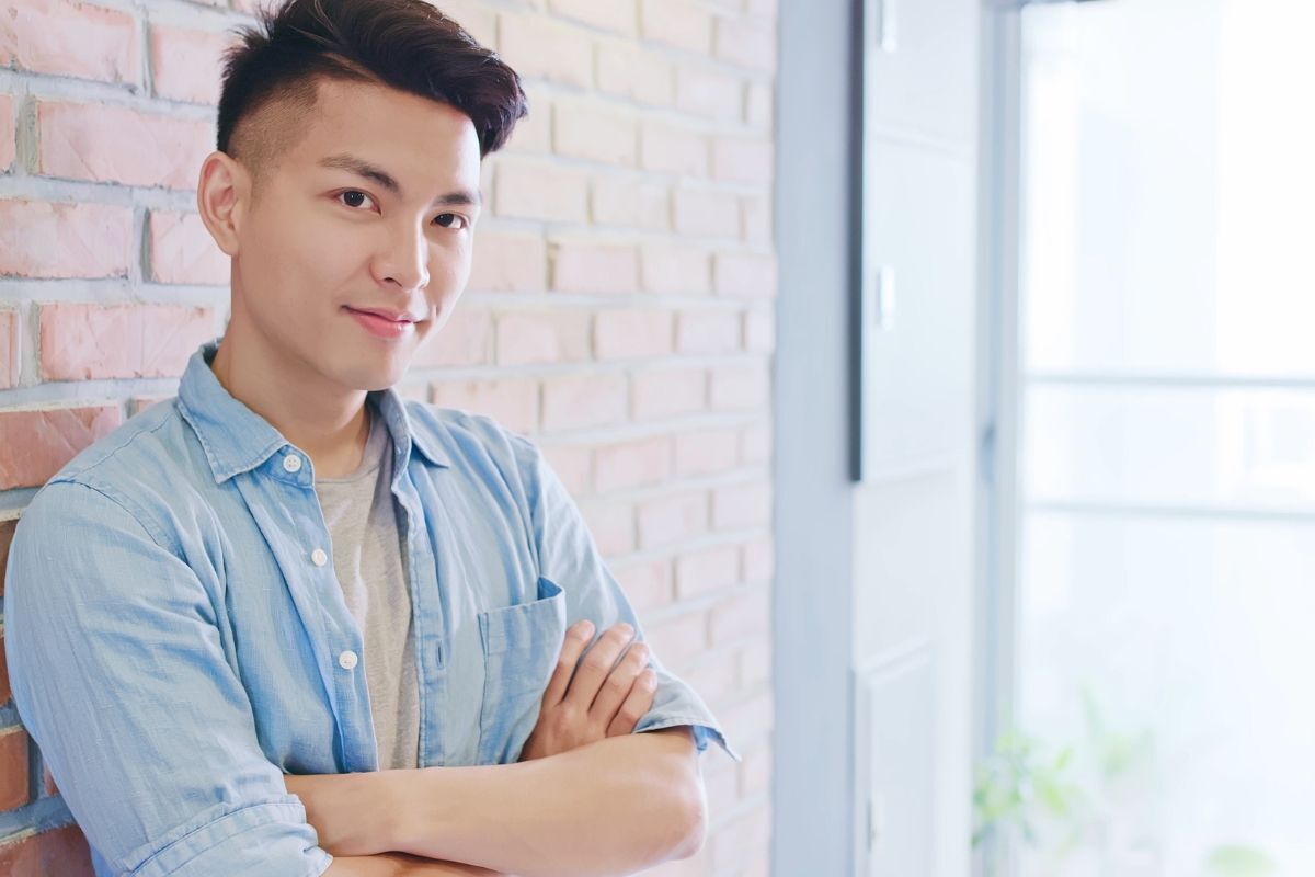tóc crop nam đẹp chàng trai mặc áo sơ mi màu xanh khoanh tay mỉm cười dựa tường