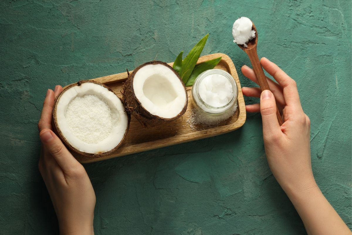 Khay gỗ màu nâu đựng hũ dầu dừa và hai nửa quả dừa khô trên nền xanh lá