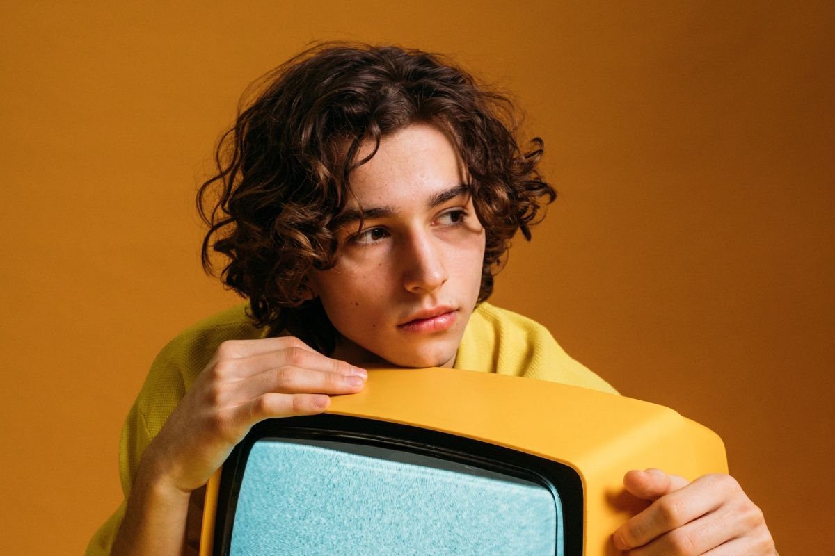 Layer xoăn rối chàng trai mặc áo len cổ lọ vàng đang ôm TV màu vàng hoài cổ