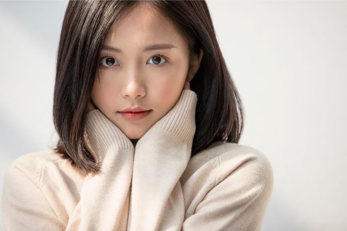 tóc mái kiểu pháp cô gái tóc bob hai mái mặc áo len mỏng màu be hai tay chạm vào cổ trên nền trắng