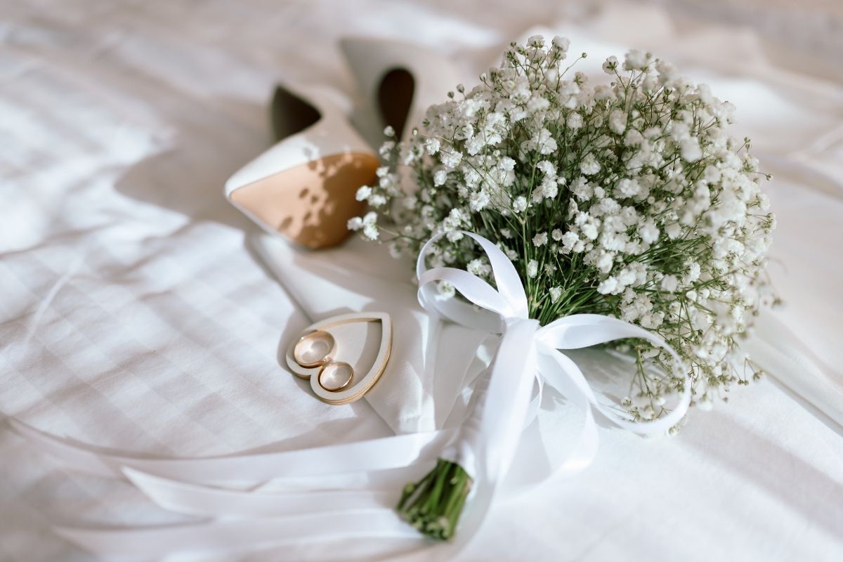 Hoa baby màu trắng buộc bằng dây ruy băng màu trắng bên cạnh cặp nhẫn cưới và đôi giày cao gót