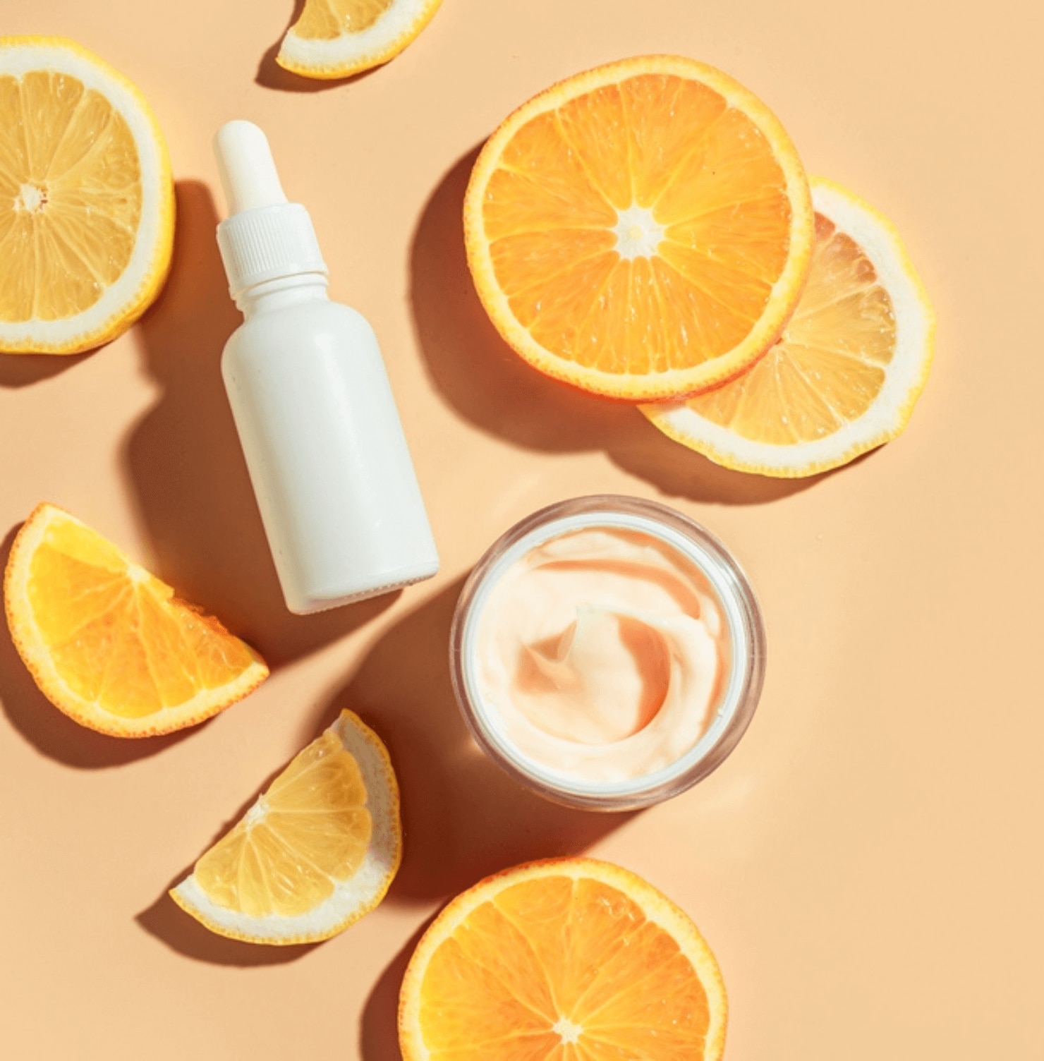 alpha arbutin kết hợp với vitamin C chai serum màu trắng và hũ kem dưỡng màu cam nhạt kế bên những lát cam trên nền cam