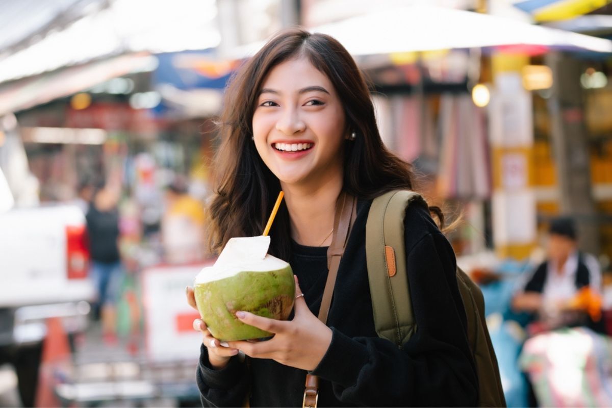 uống nước dừa mỗi ngày có đẹp da không uống nước dừa đúng cách như thế nào để làm đẹp da cô gái đeo balo cười tươi tay cầm trái dừa