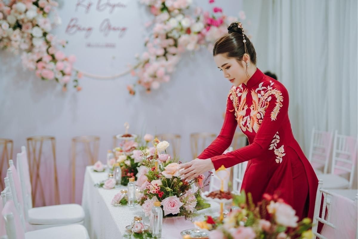Áo dài cưới màu đỏ đẹp cô dâu búi tóc đang chỉnh lại lẵng hoa trên bàn