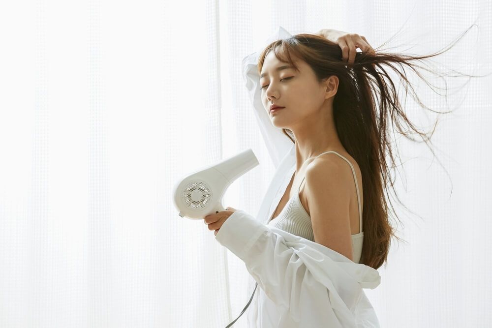 cách sấy tóc phồng không tập trung sấy khô phần chân tóc cô gái cầm máy sấy đứng bên cửa sổ màu trắng