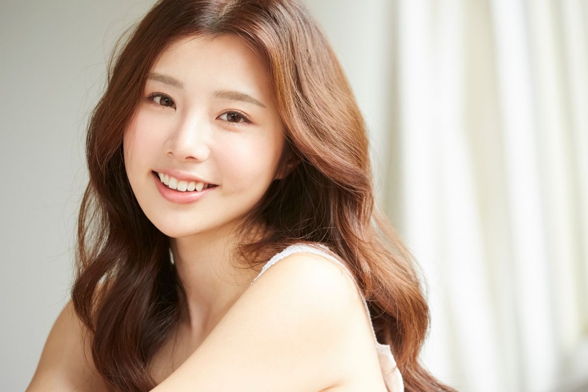 Màu tóc trầm cô gái tóc xoăn sóng nước đang nhìn vào ống kính và cười phía sau là rèm cửa màu trắng bị làm mờ