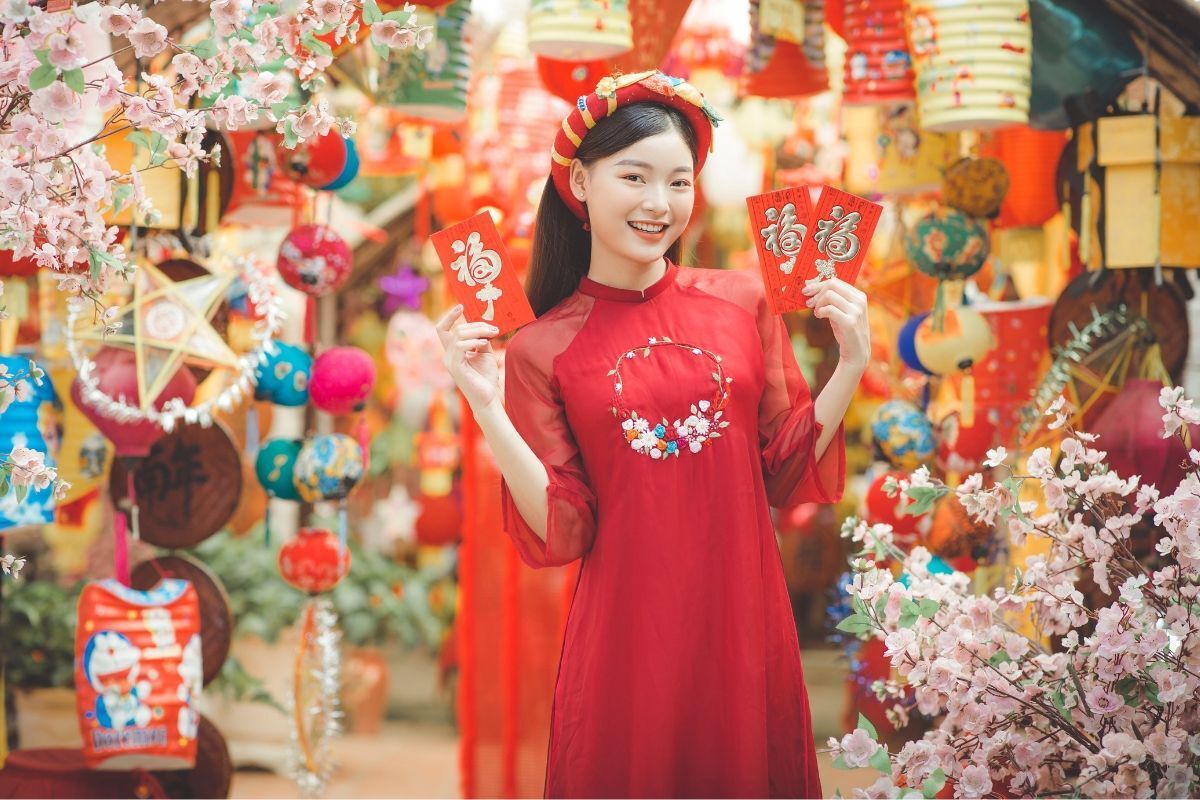 Áo dài Tết đẹp cô gái mặc áo dài màu đỏ đầu đội mấn tay cầm phong bao lì xì đỏ xung quanh là đèn lồng và hoa đào