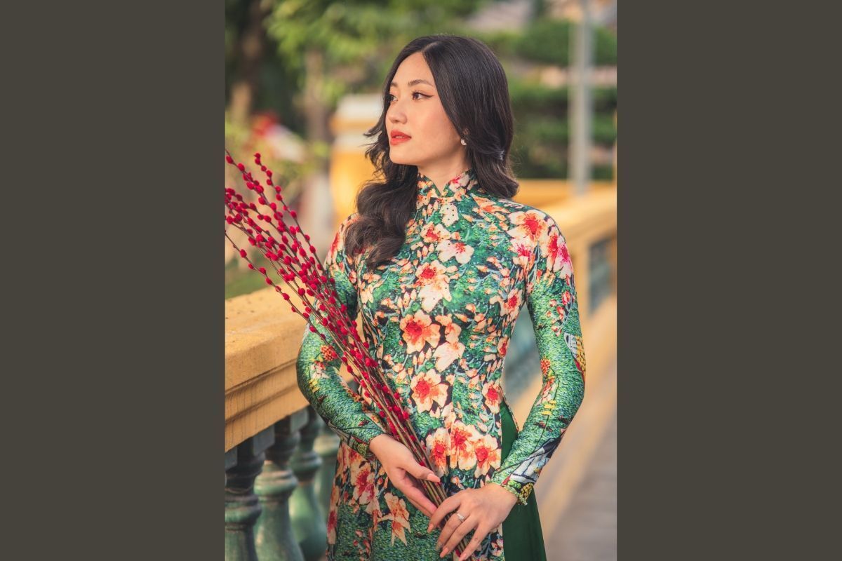 Cô gái mặc áo dài Tết màu xanh lá có hoạ tiết hoa và tay cầm bó nụ tầm xuân màu đỏ