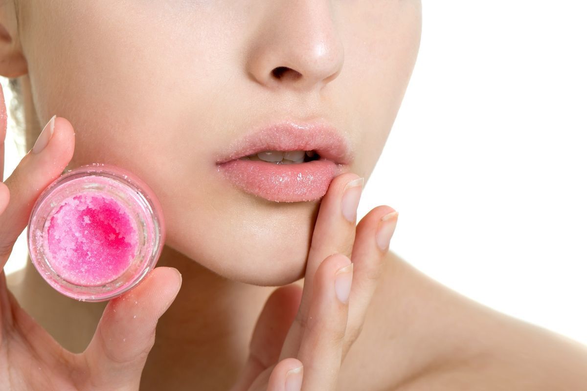 tẩy tế bào chết cho môi cô gái thoa lips scrub màu hồng lên môi taay còn lại cầm hộp scrub màu hồng trên nền trắng