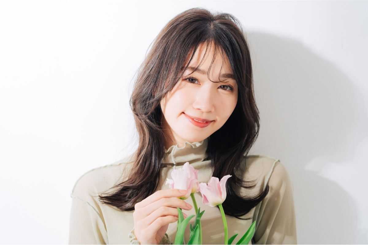 Tóc xoăn bung layer ngang vai cô gái cầm hoa màu hồng nhạt đang dựa vào tường