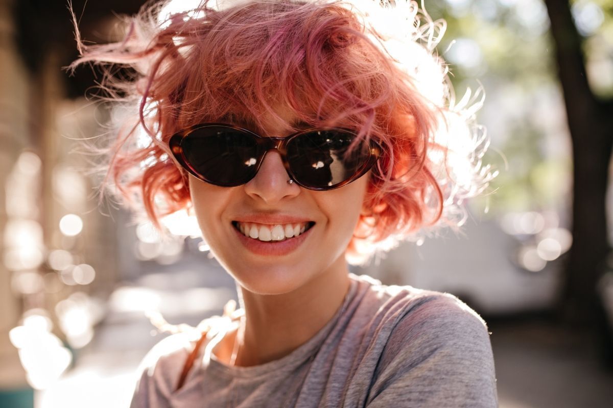 tóc nâu hồng cô gái đeo kính râm đang mỉm cười dưới ánh nắng vàng