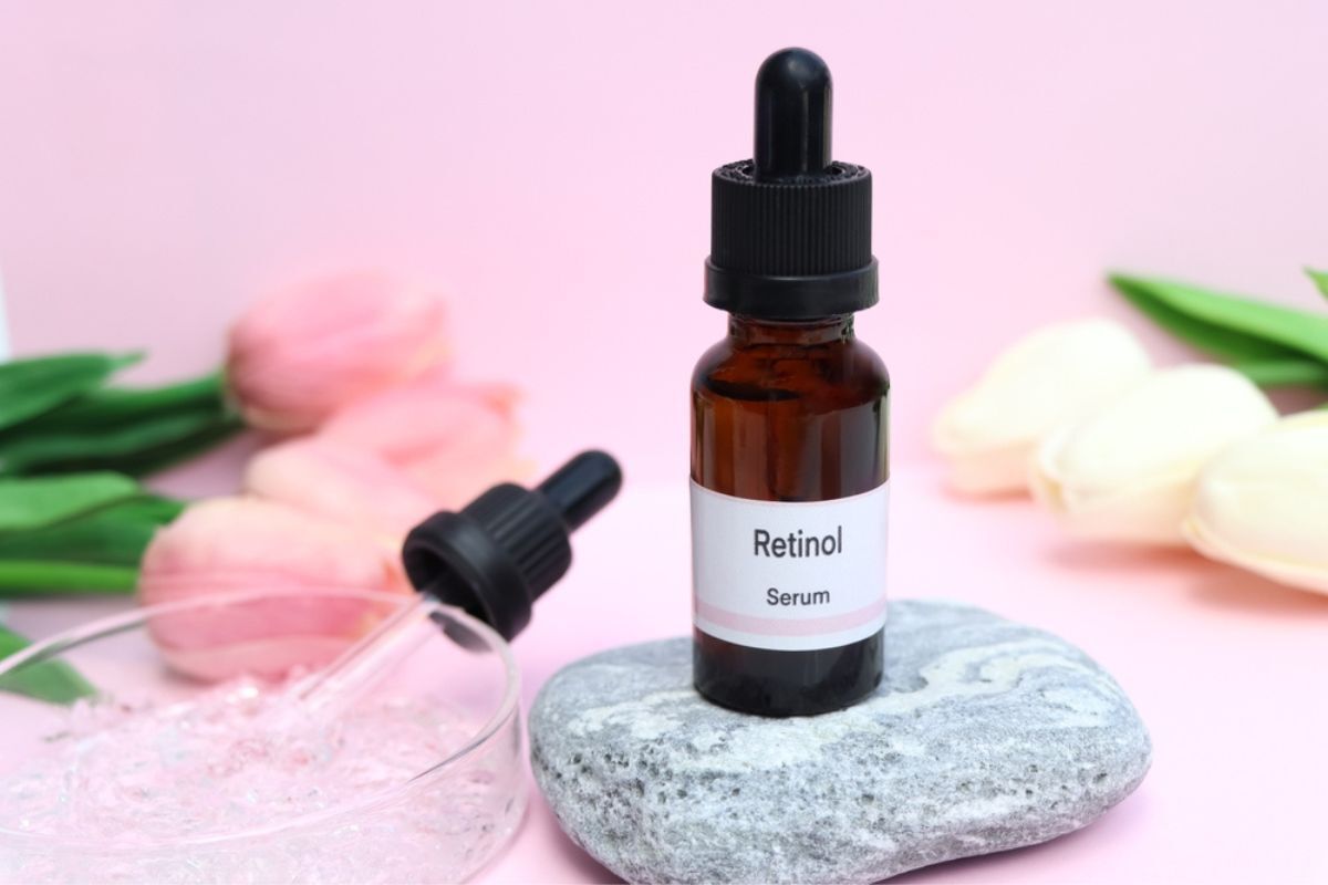 hyaluronic acid kết hợp với retinol chai serum retinol trên cục đá kế bên dĩa tròn trong suốt đằng sau là hai bó hoa tulip màu trắng và hồng trên nền hồng