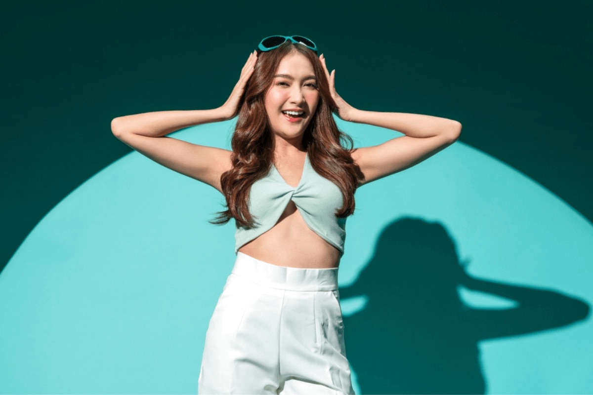 phục hồi tóc hư tổn cô gái đứng đưa tay lên đầu cười tươi ánh nắng chiếu vô in bóng trên tường màu xanh ngọc