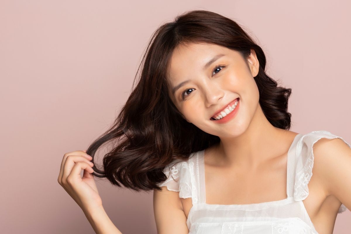 Mặt dài nên để tóc gì thử ngay kiểu tóc khắc phục khuyết điểm cô gái tóc mặc áo trắng hai dây đang mỉm cười và chạm tay vào tóc