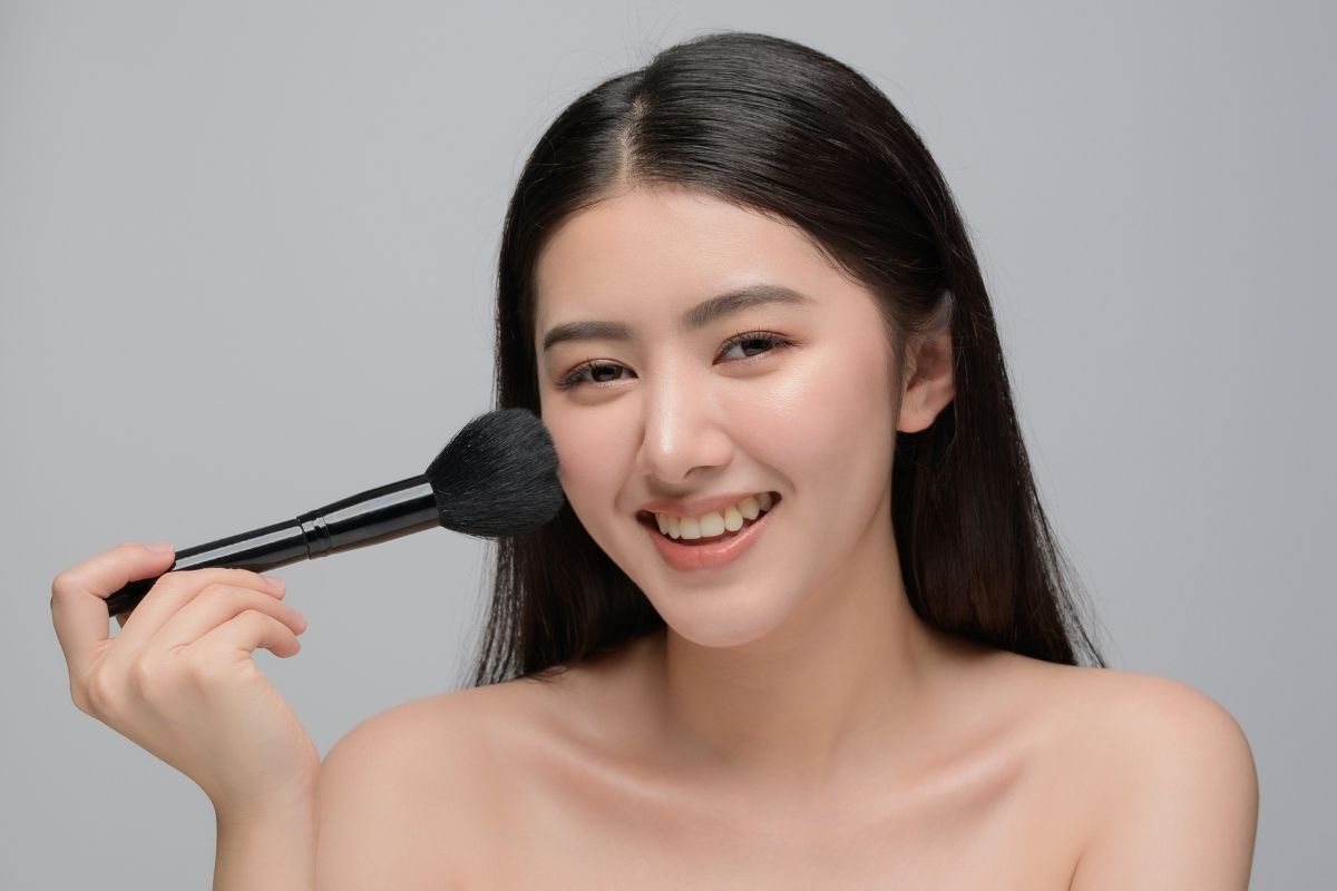 Cô gái có mái tóc đen rẽ ngôi giữa đang mỉm cười tay cầm cây cọ đánh phấn bắt sáng