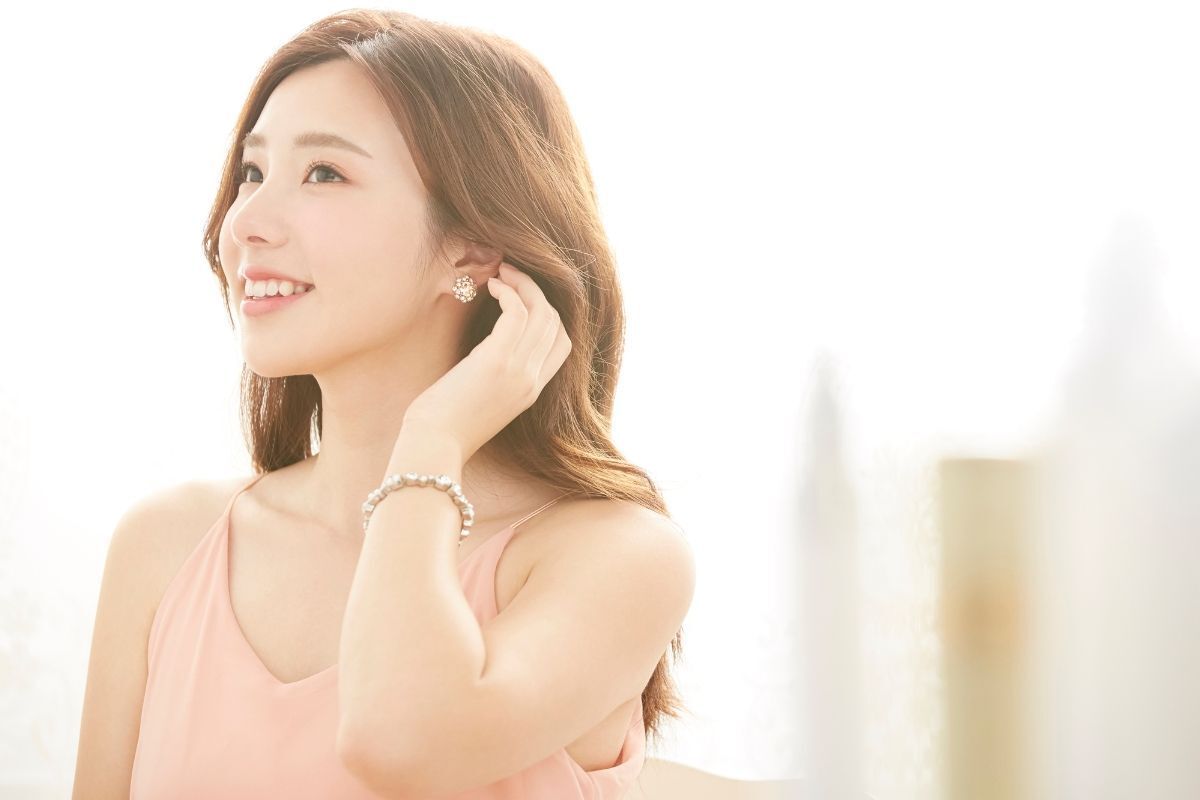 cô gái tóc màu nâu tây sáng uốn bồng bềnh mặc áo dây màu hồng nhạt nhìn nghiêng cười tay trái đang vén tóc trong căn phòng ngập nắng