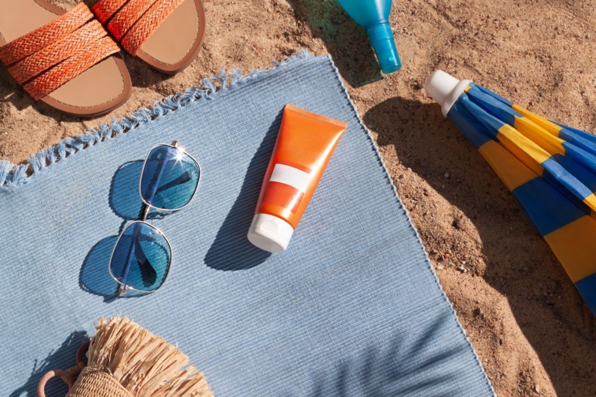 tắm nắng tắm nắng đúng cách cho người lớn mắt kính mát tuýp kem chống nắng trên khăn xanh trải trên cát kế bên cây dù và đôi dép đi biển