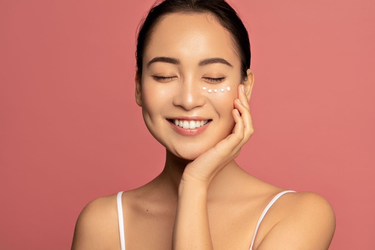 kem xóa nhăn vùng mắt cô gái thoa kem dưới vùng mắt cười tươi nhắm mắt trên nền hồng