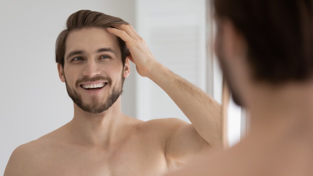 dưỡng tóc nam cách làm tóc mềm mượt cho nam chàng trai nhìn vào gương lấy tay luồn vào tóc cười tươi